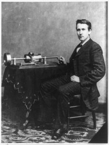 Thomas Edison is sitting in a chair beside his phonograph.
