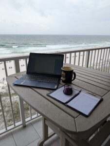 A laptop, a cup of coffee, and a notepad on a table overlooking the water.